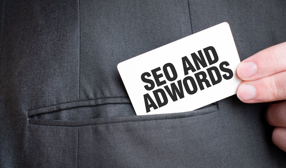 Hand holding a card that reads SEO and AdWords near a suit pocket, representing SEO vs paid ads strategies.